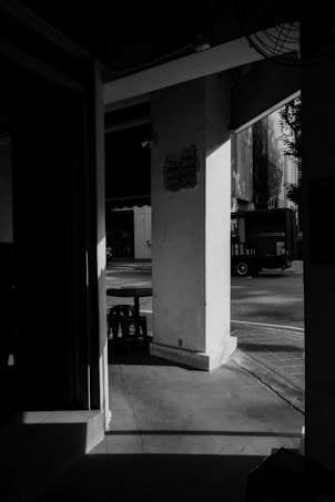 A monochrome view of an urban setting with strong contrasts of light and shadow. A structural column forms the central focus, casting shadows on a tiled walkway. In the background, a street is visible with a vehicle parked and an industrial building. The image portrays a quiet and somewhat empty scene.