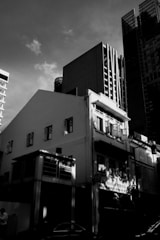 Urban scene featuring a mix of modern and traditional architecture with high-rise buildings and a smaller, older structure in the foreground. The play of light and shadow adds depth, with the sun illuminating parts of the buildings. The sky is partly cloudy, creating a dramatic effect.