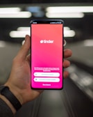A hand holding a smartphone displaying the Tinder app login screen, with options to log in using Google or a phone number. The background is blurred, suggesting a dynamic setting, possibly a moving walkway or escalator.