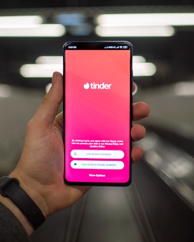 A hand holding a smartphone displaying the Tinder app login screen, with options to log in using Google or a phone number. The background is blurred, suggesting a dynamic setting, possibly a moving walkway or escalator.