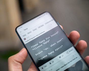 A hand is holding a smartphone displaying the 9gag app with a humorous text post about using the app repeatedly. The app's interface includes tabs and options for memes and funny content.