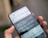 A hand is holding a smartphone displaying the 9gag app with a humorous text post about using the app repeatedly. The app's interface includes tabs and options for memes and funny content.