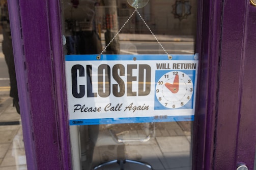 A sign hanging on a glass door indicates that a shop is closed. The sign features a clock with movable dials showing when the shop will return to being open. The frame is a deep purple, and there is a street and some reflections visible in the background.