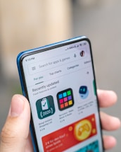 A hand is holding a smartphone displaying an app store interface. The screen shows recently updated apps with icons and ratings. The top bar includes a search function and navigation tabs.