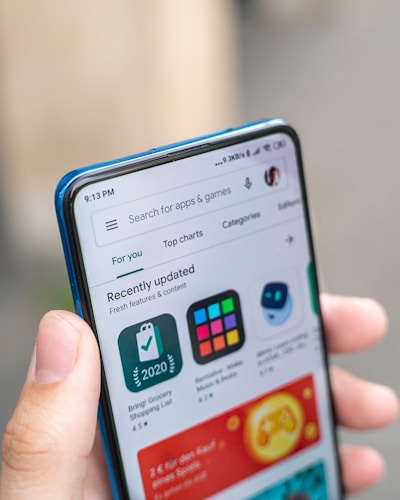 A hand is holding a smartphone displaying an app store interface. The screen shows recently updated apps with icons and ratings. The top bar includes a search function and navigation tabs.