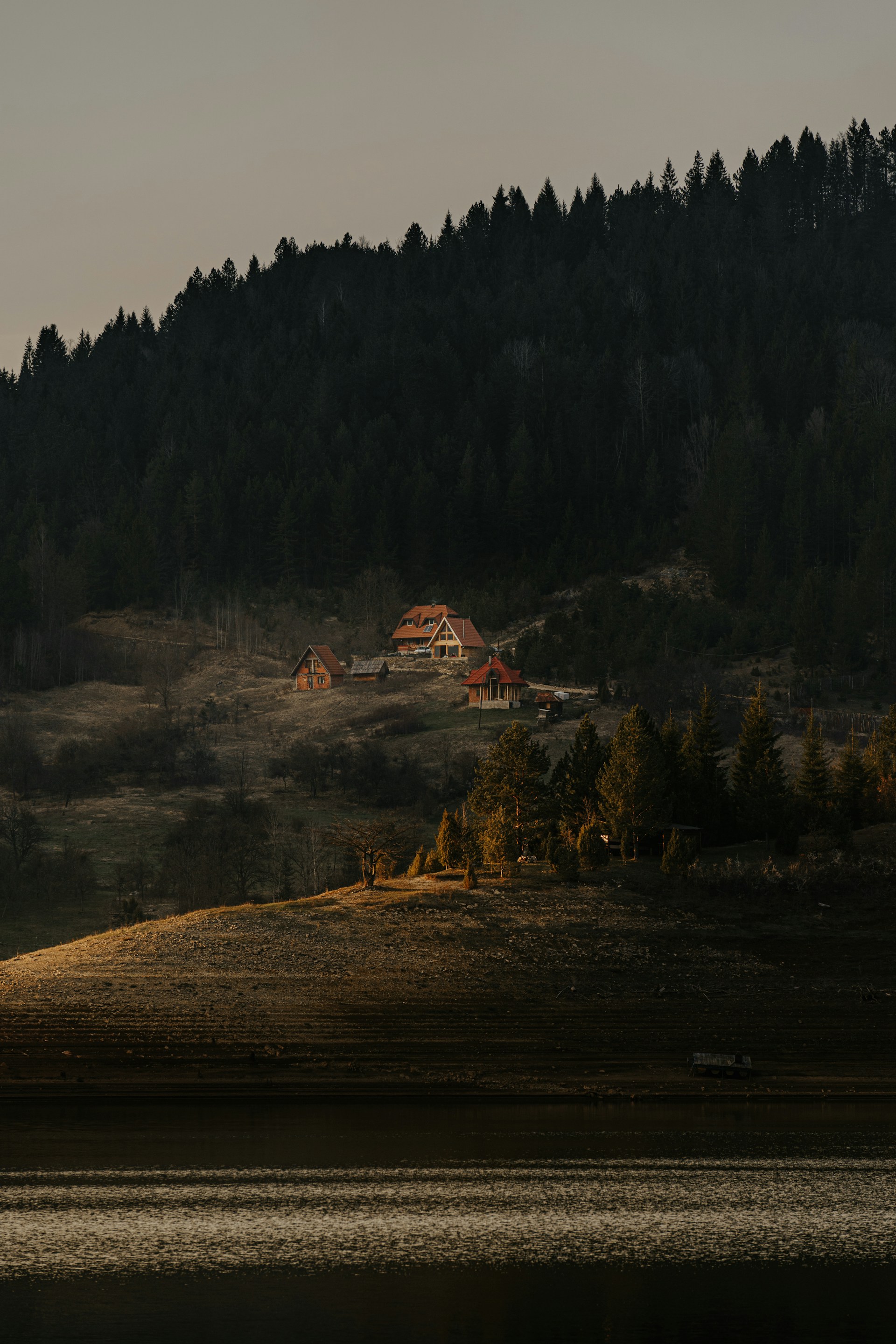 Several cabins with pitched roofs are nestled on a hillside surrounded by dense forest. The area is bathed in soft, warm sunlight, creating a tranquil and secluded atmosphere. A lake reflects the light, adding to the serene setting.