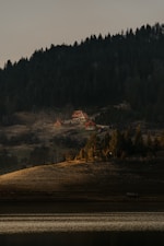 Several cabins with pitched roofs are nestled on a hillside surrounded by dense forest. The area is bathed in soft, warm sunlight, creating a tranquil and secluded atmosphere. A lake reflects the light, adding to the serene setting.