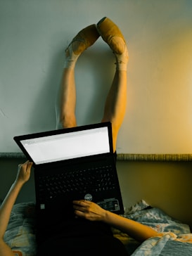 A person is lying on a bed with their legs extended upwards and resting against a wall. They are using a laptop, with the screen visible and the keyboard facing towards them. The person is wearing ballet pointe shoes on their feet.