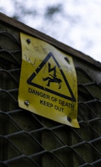 A yellow warning sign is attached to a metal fence. The sign includes a triangle symbol with a figure being struck by lightning, indicating a high voltage hazard. The text reads 'Danger of Death Keep Out'. The background is blurred, showing greenery and an overcast sky.