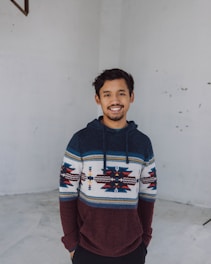 A person is standing in a minimalist indoor setting with a plain background. They are wearing a colorful, patterned hoodie with shades of dark blue, maroon, and white. The person has a cheerful expression and is looking directly at the camera.