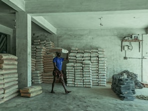 A person is carrying a bag of cement on their shoulder inside a storage room. The room contains several stacks of cement bags and a pile of metal rods or wire. The walls and floor are concrete, and there is minimal lighting with an exposed light fixture hanging from the ceiling.