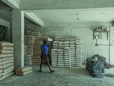 A person is carrying a bag of cement on their shoulder inside a storage room. The room contains several stacks of cement bags and a pile of metal rods or wire. The walls and floor are concrete, and there is minimal lighting with an exposed light fixture hanging from the ceiling.