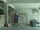 A person is carrying a bag of cement on their shoulder inside a storage room. The room contains several stacks of cement bags and a pile of metal rods or wire. The walls and floor are concrete, and there is minimal lighting with an exposed light fixture hanging from the ceiling.
