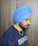 A person wearing a blue turban and a dark blue t-shirt with white text. The person has a beard and a gold chain necklace. The background is a neutral colored wall.