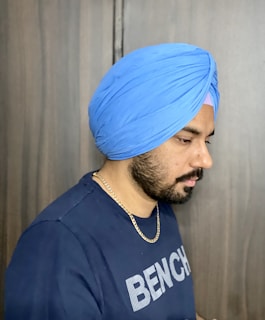 A person wearing a blue turban and a dark blue t-shirt with white text. The person has a beard and a gold chain necklace. The background is a neutral colored wall.