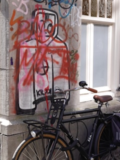 A wall covered with graffiti displaying a large, stylized figure in bold outlines and predominantly red accents. A black bicycle with a brown seat is parked against the graffiti-covered wall. The bike features a front light and a rear rack. The scene is set next to a window with white framing, casting shadows on the graffiti.