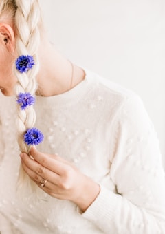 A person with long blond hair styled in a braid decorated with vibrant blue flowers. The individual is wearing a white sweater with textured patterns and holding a section of their hair. Light and soft colors dominate the image, creating a serene and delicate atmosphere.