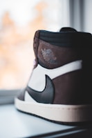 A close-up of a high-top sneaker featuring a sleek design with a combination of brown, black, and white colors. The shoe displays a winged logo with the words 'Air Jordan' embossed on the upper part. It is positioned near a window with soft natural light highlighting its details.