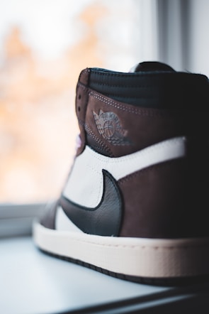 A close-up of a high-top sneaker featuring a sleek design with a combination of brown, black, and white colors. The shoe displays a winged logo with the words 'Air Jordan' embossed on the upper part. It is positioned near a window with soft natural light highlighting its details.
