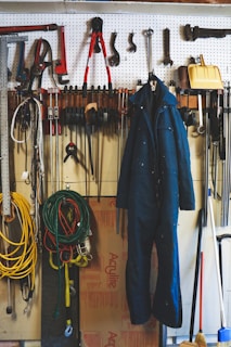 A neatly organized garage workspace featuring a pegboard mounted on the wall with an array of tools including hammers, wrenches, pliers, and clamps. A blue work jumpsuit hangs prominently on the pegboard. Below the board, coiled extension cords and ropes are visible, along with a broom and a yellow snow shovel. The background includes a partially visible cardboard box and some additional workshop items.
