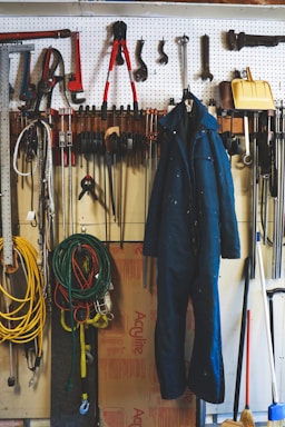 A neatly organized garage workspace featuring a pegboard mounted on the wall with an array of tools including hammers, wrenches, pliers, and clamps. A blue work jumpsuit hangs prominently on the pegboard. Below the board, coiled extension cords and ropes are visible, along with a broom and a yellow snow shovel. The background includes a partially visible cardboard box and some additional workshop items.