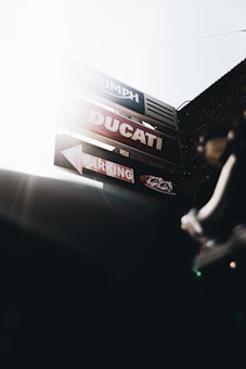 A set of signage featuring the brands Triumph and Ducati is mounted against a brick wall with a bright, sunlit sky in the background. The signs include bold text and arrows indicating parking directions. The image is partially obscured by shadow.
