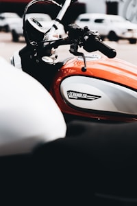 A close-up view of a motorcycle displaying the fuel tank and handlebars. The tank features a logo that reads 'Scrambler Ducati.' The surrounding environment suggests an outdoor setting with blurred vehicles in the background.
