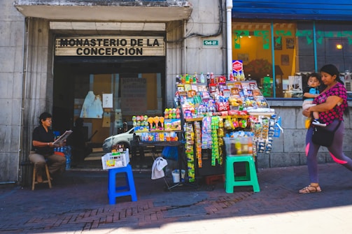 An outdoor street scene features a small convenience stall laden with a variety of snacks, drinks, and small items. A woman holding a child walks past the bustling stand. To the left, a person is seated on a stool reading a newspaper in front of a building labeled 'Monasterio de la Concepción'. The scene captures a busy urban environment with a mix of activity.