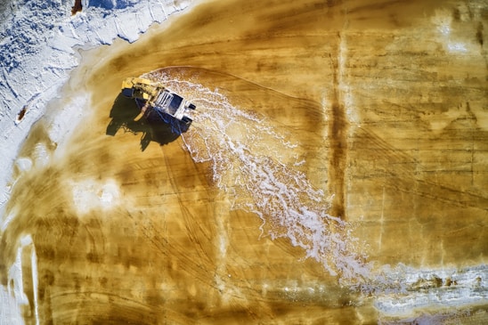 An aerial view of a large, yellow mining dump truck driving through a muddy terrain, leaving trails of water and mud in its wake. The ground is predominantly a golden brown color, indicating a possibly industrial or construction site with patches of white that could be salt or residue.