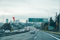 A busy urban highway is lined with traffic heading towards the city skyline, which features modern high-rise buildings. Overhead road signs direct drivers towards various destinations. The sky is overcast, and the road is bordered by trees and lamp posts.