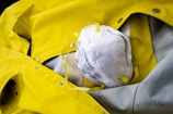 A white face mask with yellow straps lies on top of a bright yellow raincoat. The raincoat appears to be made of a waterproof material with metallic snaps and stitching.