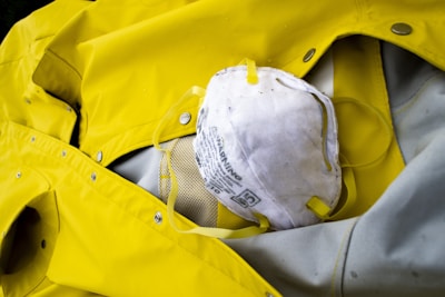 A white face mask with yellow straps lies on top of a bright yellow raincoat. The raincoat appears to be made of a waterproof material with metallic snaps and stitching.