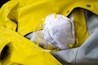 A white face mask with yellow straps lies on top of a bright yellow raincoat. The raincoat appears to be made of a waterproof material with metallic snaps and stitching.
