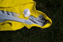 A bright yellow raincoat lies on grass, with a white face mask positioned on top of it. The raincoat is spread out, showing its inside lining, and the grass provides a contrasting natural background.