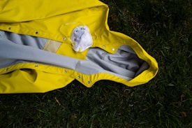 A bright yellow raincoat lies on grass, with a white face mask positioned on top of it. The raincoat is spread out, showing its inside lining, and the grass provides a contrasting natural background.