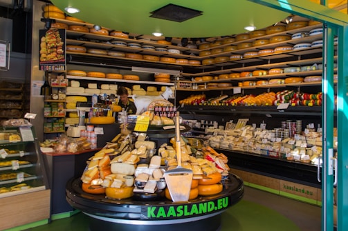 A cheese shop filled with various types of cheeses displayed on shelves and counters. Large wheels of cheese are stacked on the shelves along the walls, while smaller pieces are arranged on a central display table. There are signs indicating prices, and a worker is seen behind the counter assisting customers. The shop has a welcoming and abundant atmosphere.
