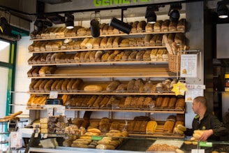 An assortment of bread is displayed on several shelves in a bakery. The shelves are stocked with various types of bread, including loaves and baguettes. A person, possibly a staff member, is visible on the right handling some bread. There are also signs and price tags visible near the bread on display.