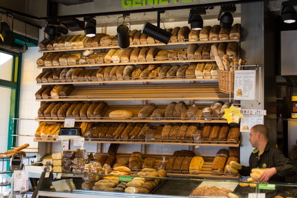 An assortment of bread is displayed on several shelves in a bakery. The shelves are stocked with various types of bread, including loaves and baguettes. A person, possibly a staff member, is visible on the right handling some bread. There are also signs and price tags visible near the bread on display.