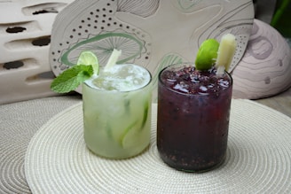 Two refreshing beverages are placed on a textured circular mat. The drink on the left is a light green cocktail garnished with fresh mint leaves and a slice of lime. The one on the right is a dark berry-colored drink with visible fruit seeds and a lime wedge. Both drinks are served in short clear glasses with ice.