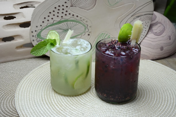 Two refreshing beverages are placed on a textured circular mat. The drink on the left is a light green cocktail garnished with fresh mint leaves and a slice of lime. The one on the right is a dark berry-colored drink with visible fruit seeds and a lime wedge. Both drinks are served in short clear glasses with ice.