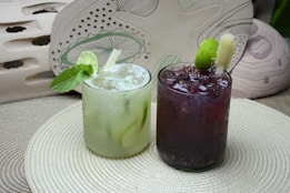 Two refreshing beverages are placed on a textured circular mat. The drink on the left is a light green cocktail garnished with fresh mint leaves and a slice of lime. The one on the right is a dark berry-colored drink with visible fruit seeds and a lime wedge. Both drinks are served in short clear glasses with ice.