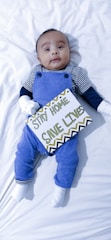 A baby lying on a white sheet wearing a striped shirt with blue overalls. The baby is holding a sign with the text 'Stay Home, Save Lives' written in green and black letters on a white background with decorative patterns.