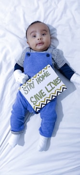 A baby lying on a white sheet wearing a striped shirt with blue overalls. The baby is holding a sign with the text 'Stay Home, Save Lives' written in green and black letters on a white background with decorative patterns.