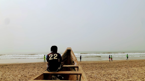 A person wearing a jersey with the number 22 is sitting on a wooden boat on a sandy beach. In the background, there are several people strolling along the shore near the waves of the ocean under a cloudy sky.