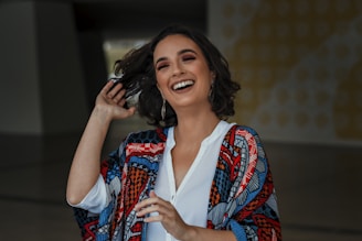 A woman with shoulder-length, wavy hair is laughing joyfully. She is wearing a colorful, patterned shawl over a white shirt and has hoop earrings. Her makeup is well-done, and she appears to be in a good mood. The background is slightly blurred and includes abstract designs.
