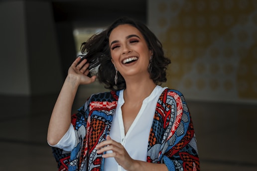 A woman with shoulder-length, wavy hair is laughing joyfully. She is wearing a colorful, patterned shawl over a white shirt and has hoop earrings. Her makeup is well-done, and she appears to be in a good mood. The background is slightly blurred and includes abstract designs.