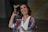 A woman with shoulder-length, wavy hair is laughing joyfully. She is wearing a colorful, patterned shawl over a white shirt and has hoop earrings. Her makeup is well-done, and she appears to be in a good mood. The background is slightly blurred and includes abstract designs.