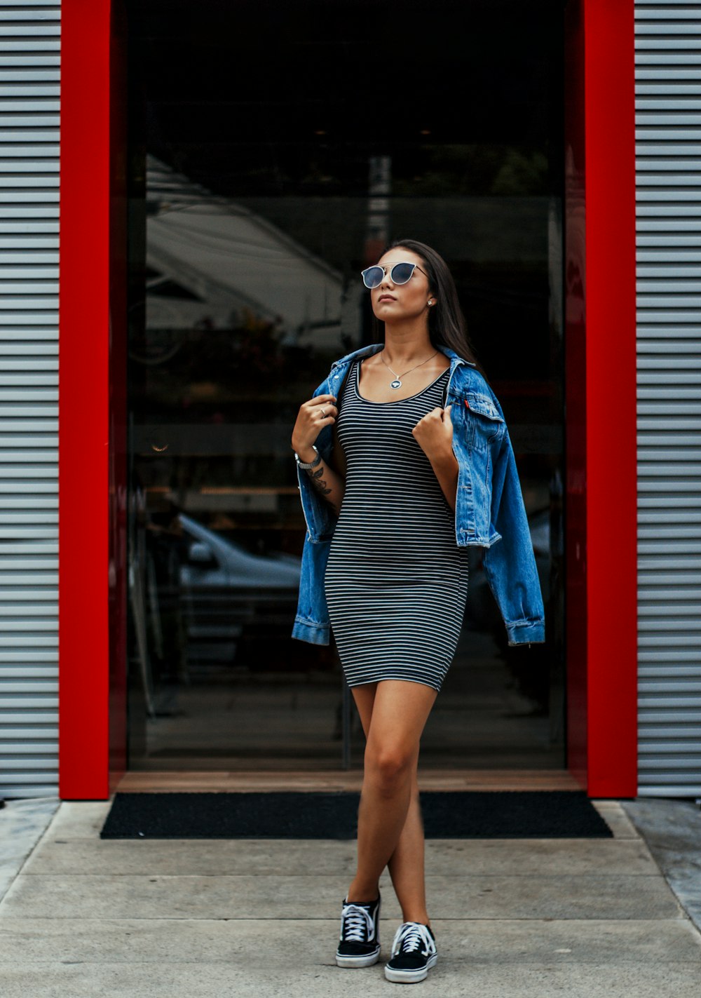 woman in blue and white stripe tank top and blue denim jacket standing beside red wall