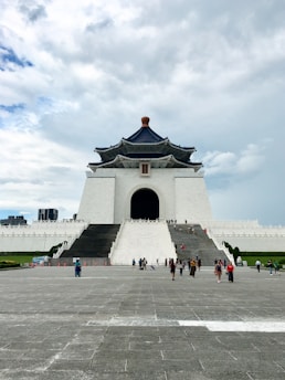A large, imposing white building with a dark blue roof and a traditional architectural design. The building is accessed by a wide set of stairs, and people are walking towards it. The sky is partly cloudy, and the surroundings are spacious and open.