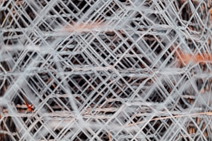 A complex pattern of interwoven metal wires creating a mesh-like structure, with a focus on the intricate crisscross and layering of the wires. The metal has a slightly worn, industrial appearance.